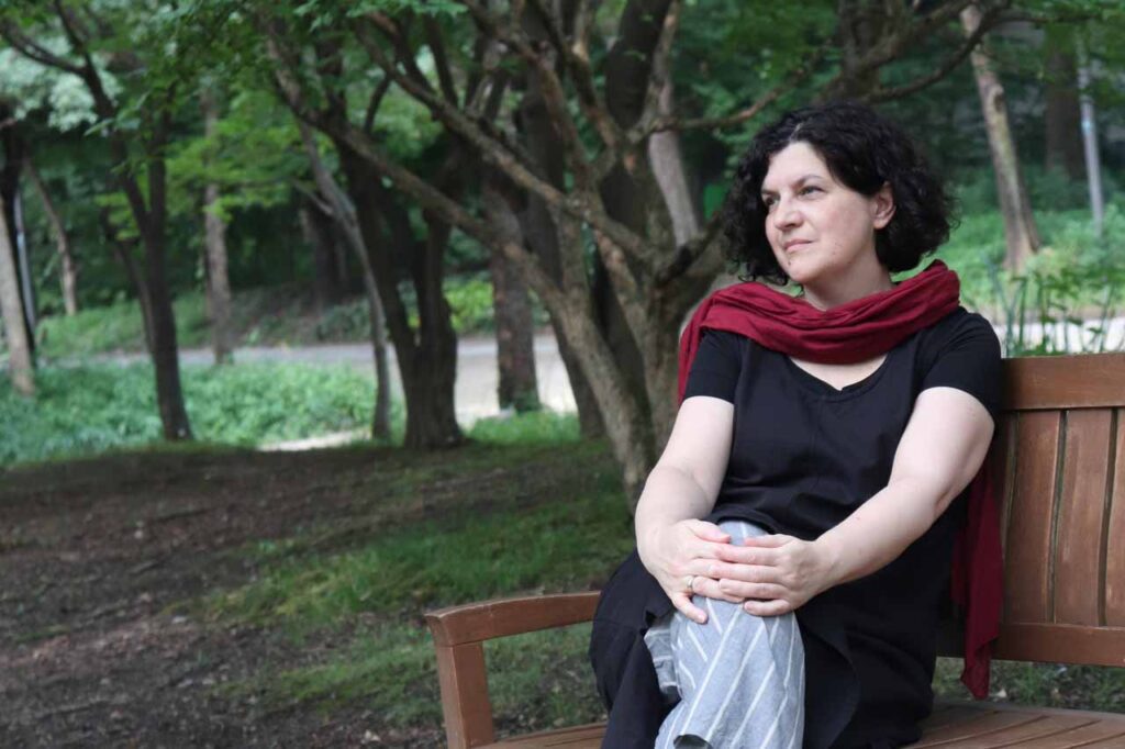 Ciemnowłosa kobieta, ubrana w czarną, długa bluzkę, szare spodnie w białe paski i bordową chustę siedzi na drewnianej ławce w parku