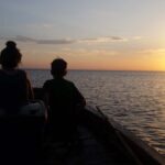 Zachód słońca na morzu, dwoje dzieci siedzi w łódce i podziwia zachód słońca.