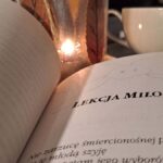 Książka otwarta na rozdziale zatytułowanym "Lekcja miłości. obok książki lampion ze świeczką i szara filizanka