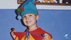 Mały, uśmiechnięty chłopiec w czerwono-żółtym stroju arlekina, w niebiesko-czerwonej czapeczce.