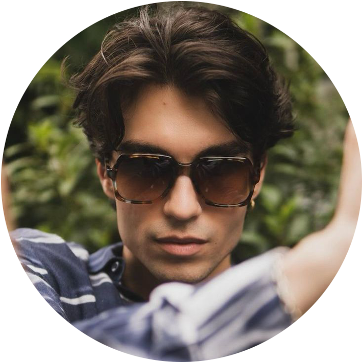 Portret młodego mężczyzny, ciemne włosy, okulary przeciwsłoneczne, fioletowo-biała bluzka, w tle zielone liście