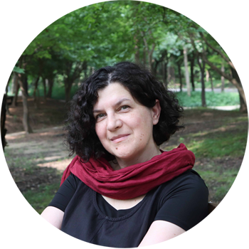 Zdjęcie portretowe, uśmiechnięta kobieta, ciemne włosy, ubrana w czarną bluzkę i bordową chustę; w tle zielone drzewa