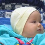 Zdjęcie portretowe małego bobaska siedzącego w wózku. Ubrany w jasnozieloną kurteczkę i żółtą, pastelową czapeczkę.