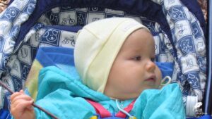 Zdjęcie portretowe małego bobaska siedzącego w wózku. Ubrany w jasnozieloną kurteczkę i żółtą, pastelową czapeczkę.