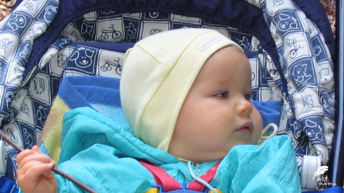 Zdjęcie portretowe małego bobaska siedzącego w wózku. Ubrany w jasnozieloną kurteczkę i żółtą, pastelową czapeczkę.