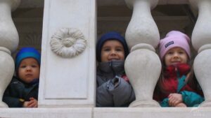 Troje uśmiechniętych dzieci wyglądających zza marmurowych tralek na schodach.