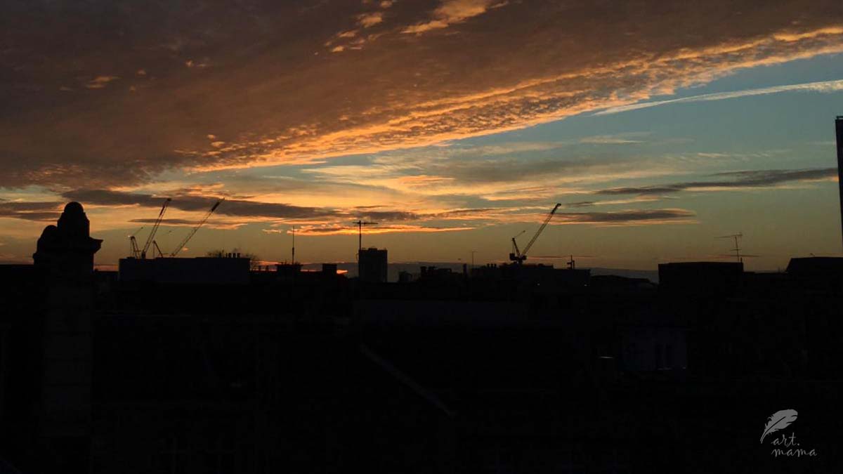 Widok miasta przy zachodzącym słońcu. Budynki całkiem ciemne, wręcz czarne, niebo z chmurami w kolorach niebieskim i różnych odcieni koloru pomarańczowego.