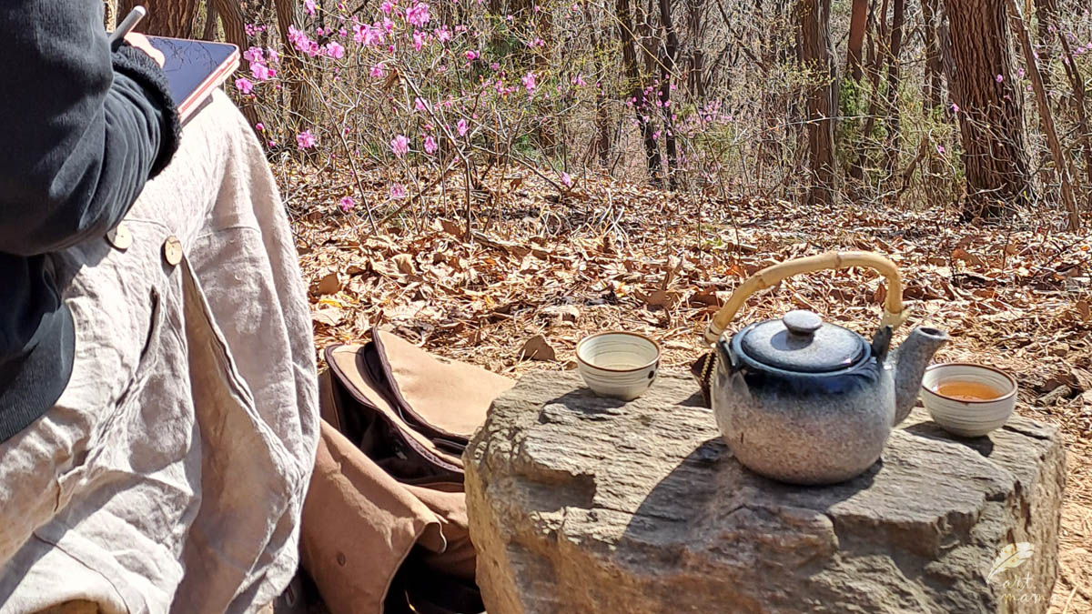 Ktoś siedzi na kamieniu w lesie (nie widać dokładnie postaci), obok na drugim kamieniu szary dzbanek z herbatą i dwie czarki; na ziemi jeszcze uschłe liście - pozostałość jesieni, ale w oddali już rozkwitają filetowe, wiosenne kwiaty